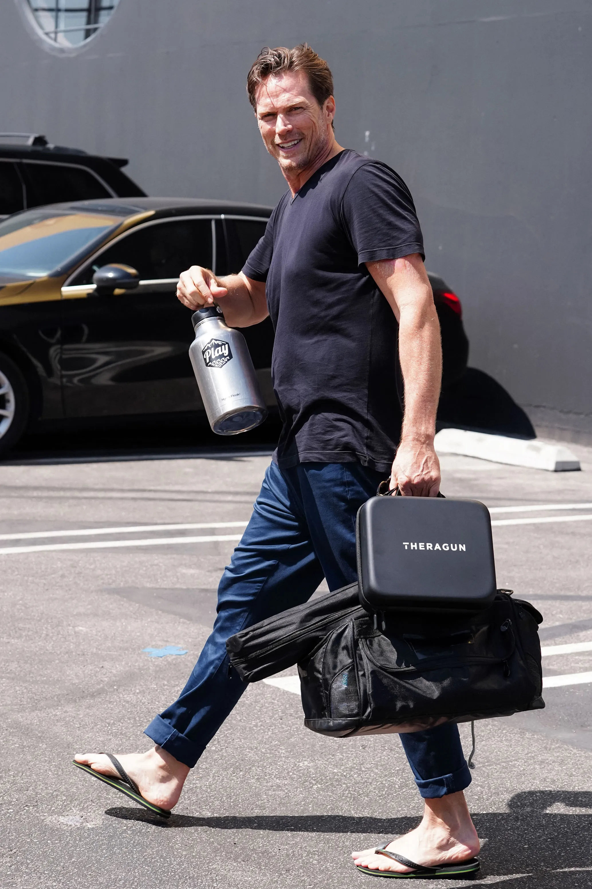 Jason Lewis walking and smiling while carrying a water bottle, a Theragun case, and a duffle bag.