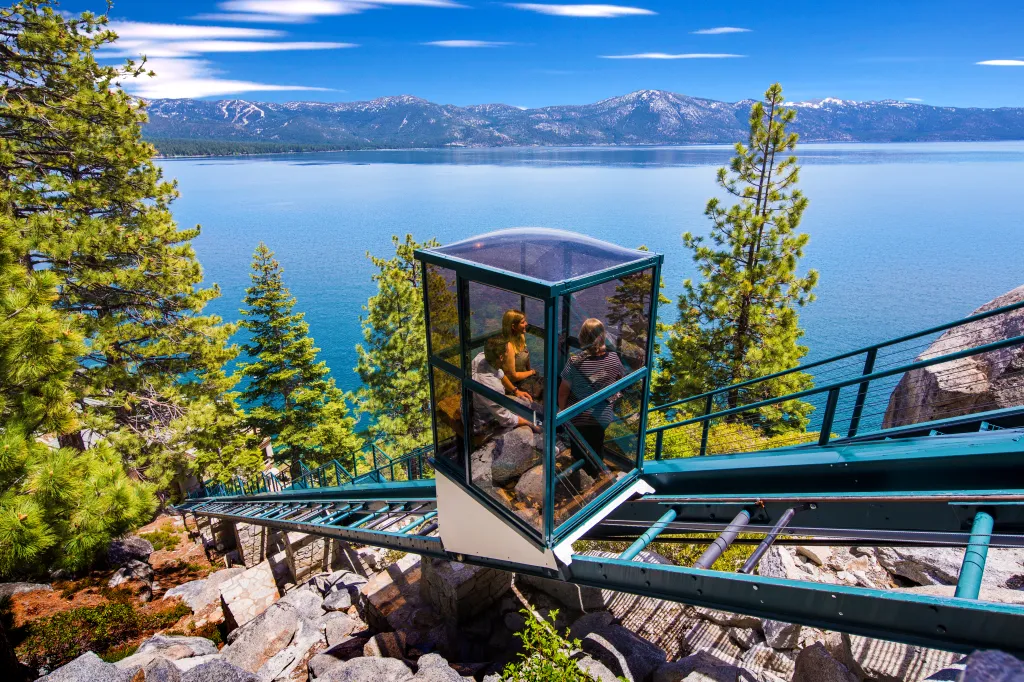 A glass funicular with three passengers descends a rocky slope toward Lake Tahoe, with pine trees in the foreground and snowy mountains in the background.