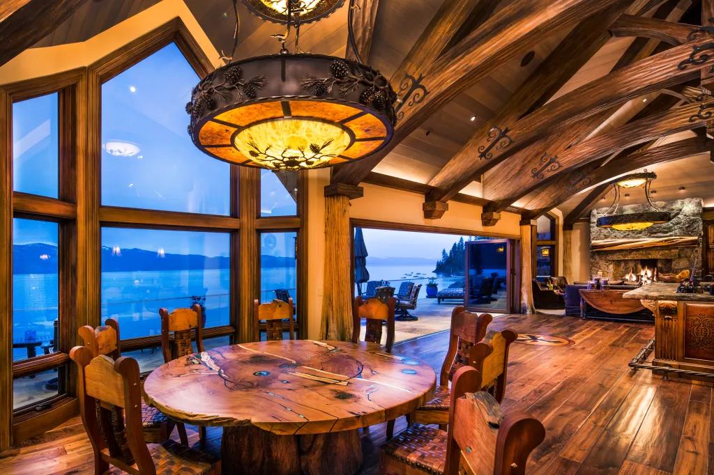 The interior of Lake Tahoe's most expensive home, featuring a large, round wooden dining table, wooden chairs, and panoramic windows overlooking the lake.