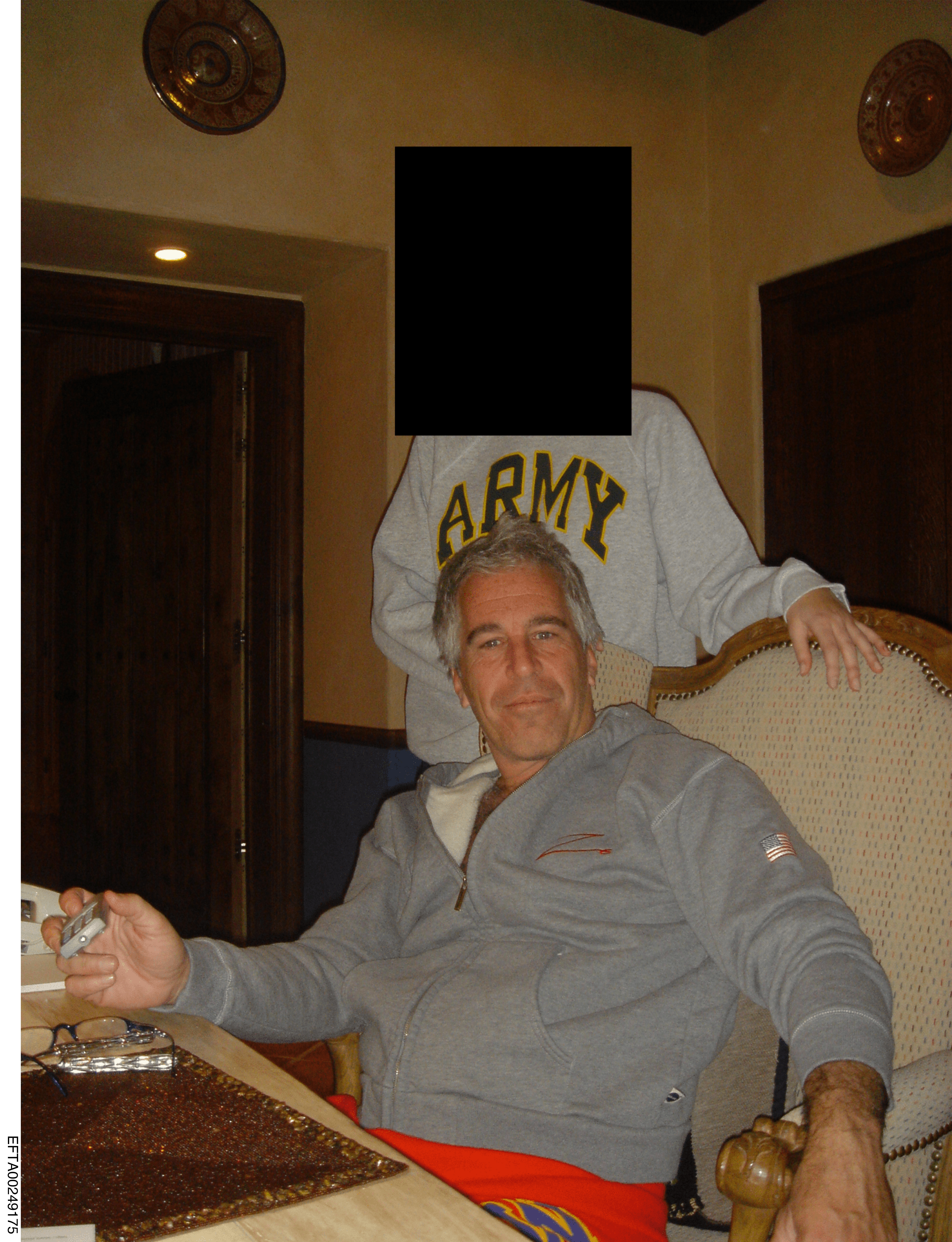 Jeffrey Epstein seated at a table, with another individual (face blacked out) standing behind him wearing an 