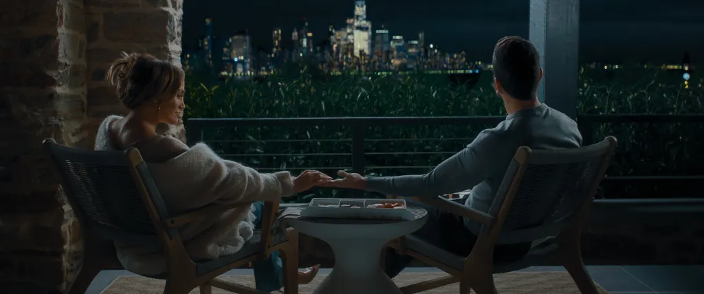 Jennifer Lopez and Brett Goldstein holding hands on a balcony at night, overlooking a city skyline.