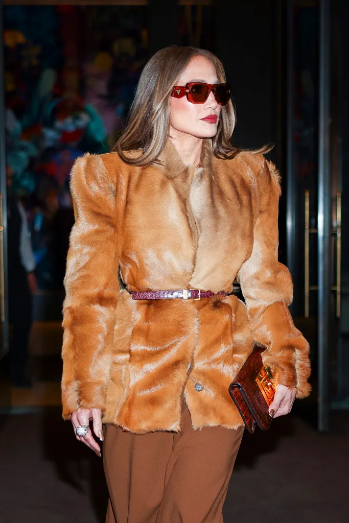 Jennifer Lopez wearing a brown faux fur coat, brown pants, red sunglasses, and carrying a brown clutch.