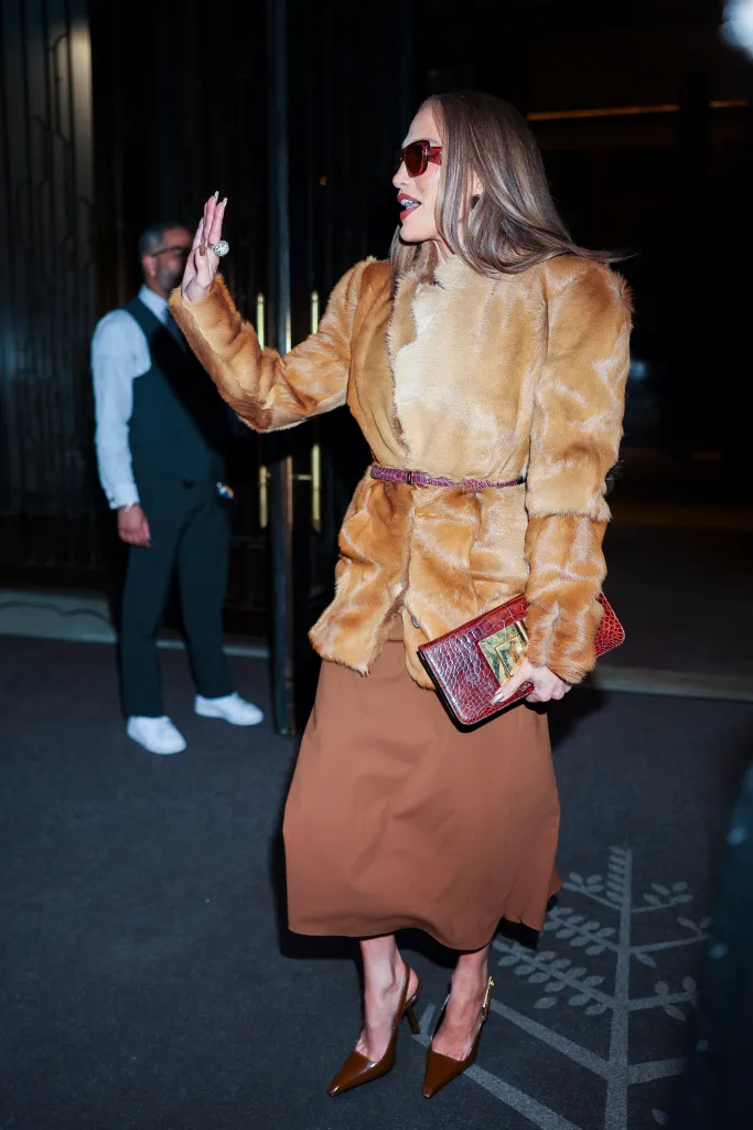 Jennifer Lopez in a fur coat, brown skirt, and sunglasses, holding a clutch and waving.