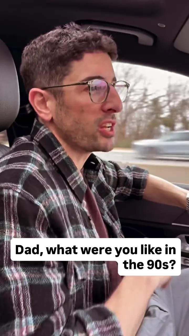 Jason Biggs driving a car, wearing a plaid shirt and glasses, with the text 