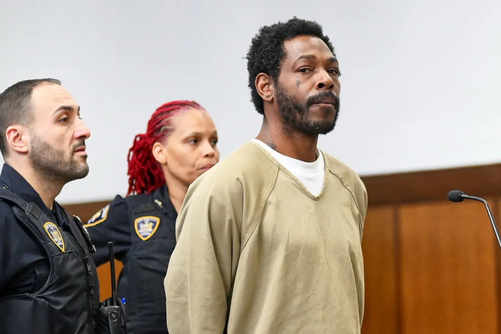 Jermaine Longmire in a tan jumpsuit, escorted by two police officers, one with red dreadlocks, in a courtroom.