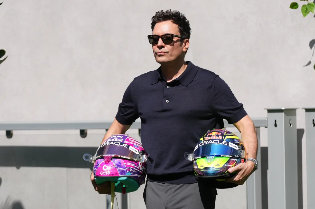 Jimmy Fallon poses with two colorful F1 racing helmets.