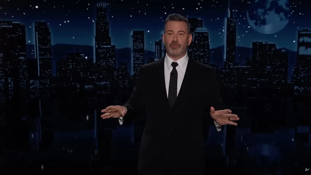 Jimmy Kimmel on stage in a suit with a city skyline and moon behind him.