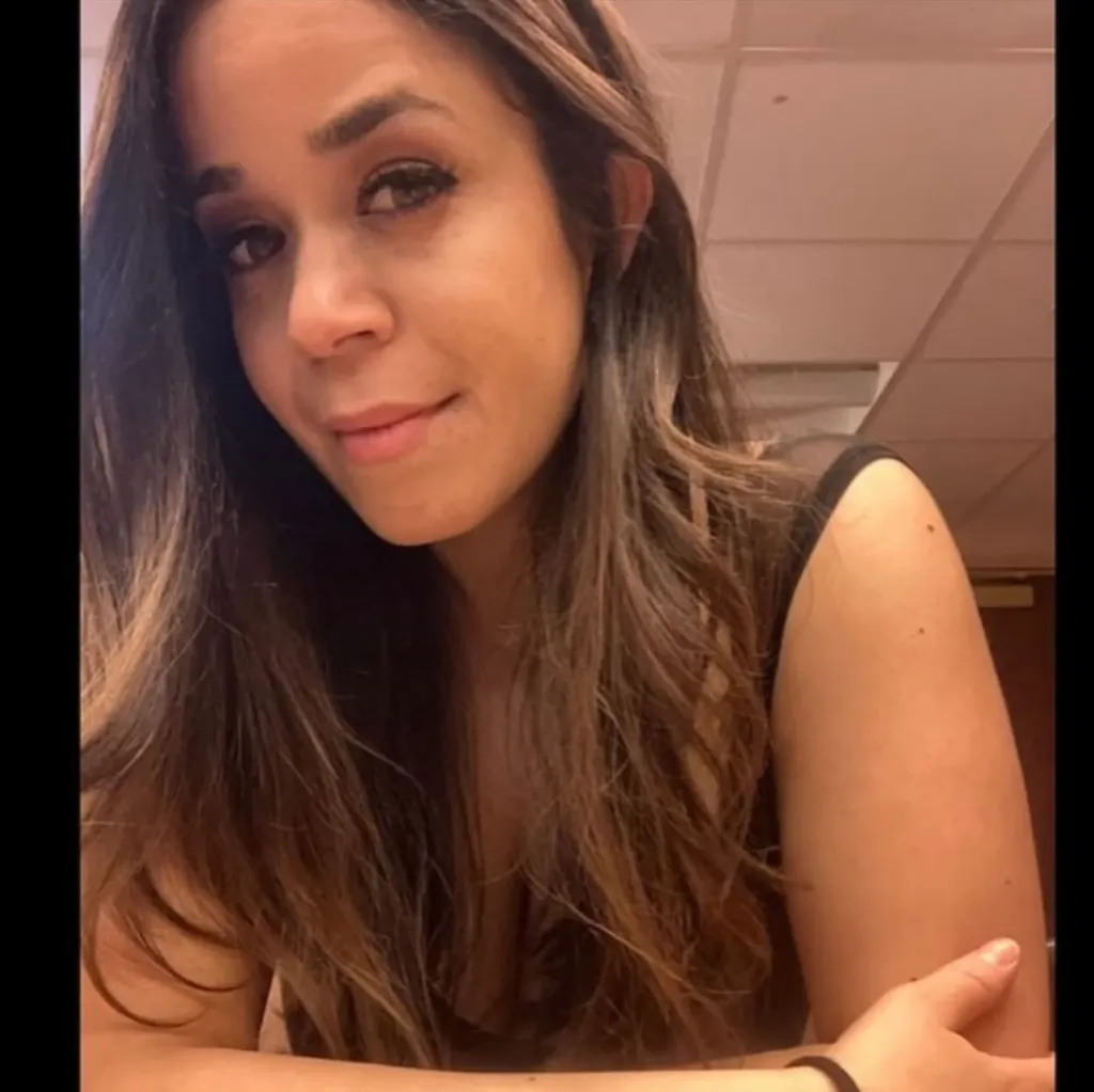 A woman with long brown hair and a dark top looks towards the camera.