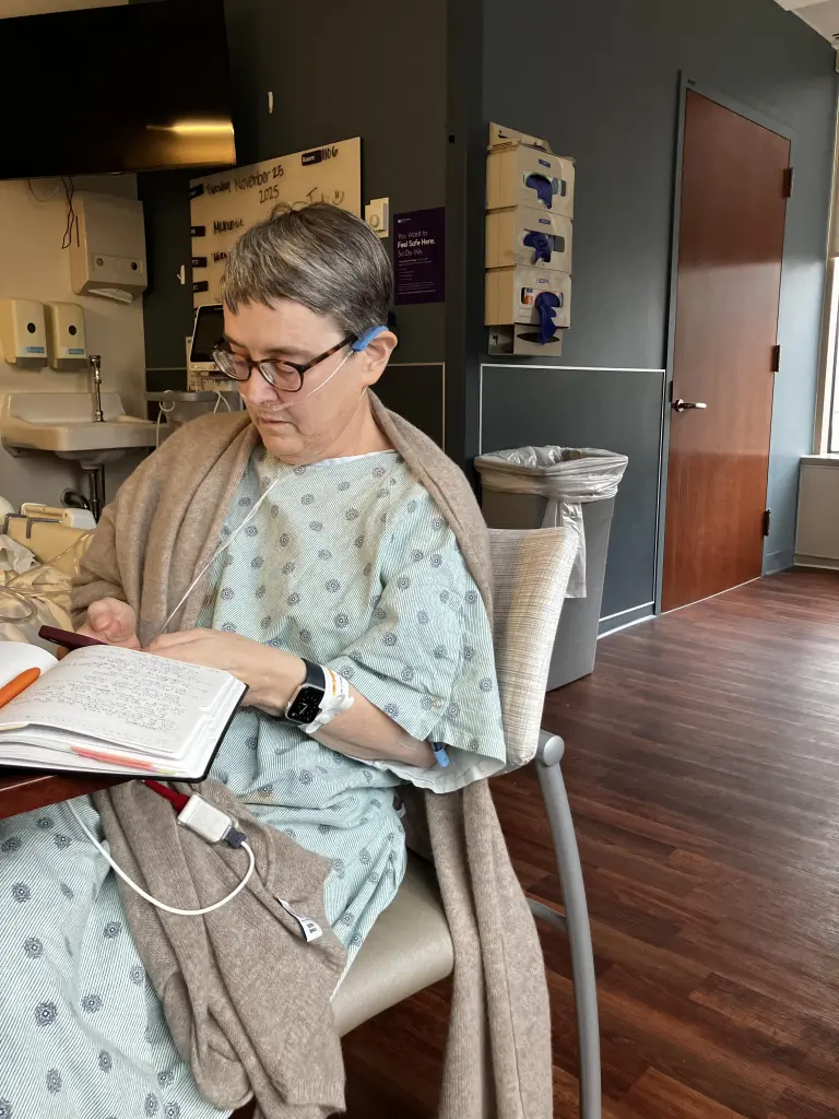 Jodi Graf, wearing a hospital gown and oxygen, writes in a notebook in her hospital room.