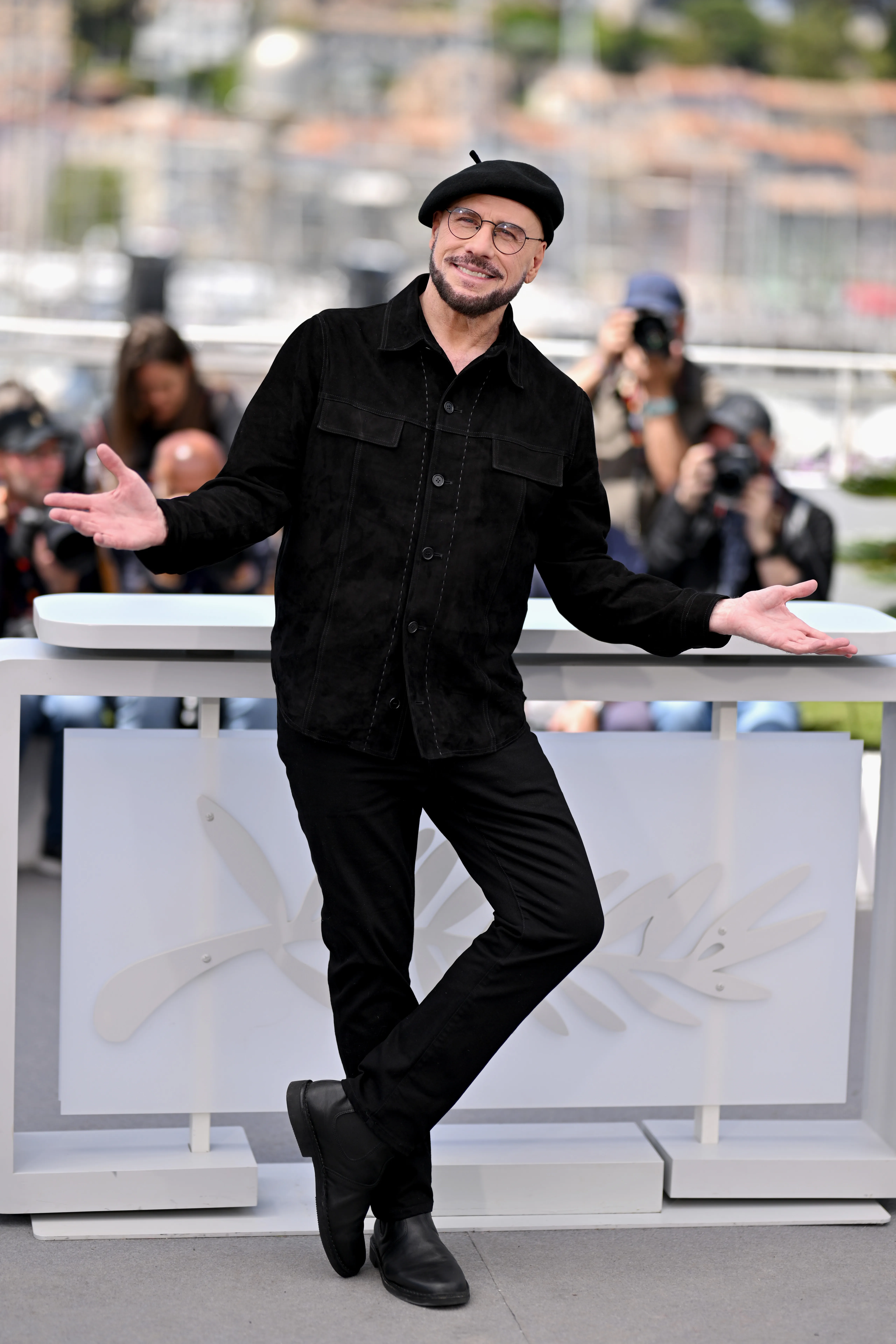 Actor John Travolta in a white beret, light pink tie, and black suit, pointing upwards at the 79th Cannes Film Festival.