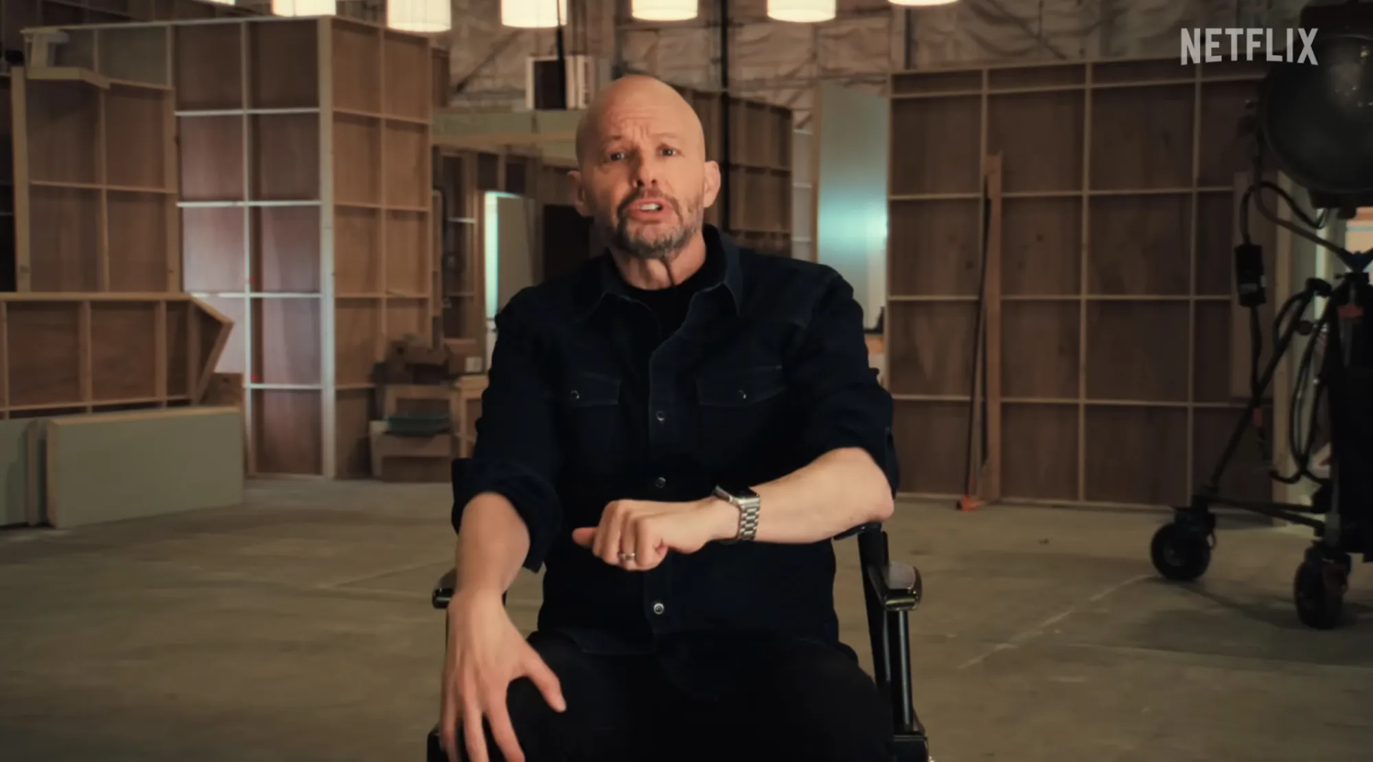 Jon Cryer sitting in a director's chair in a film set, wearing a black shirt and black pants, speaking to the camera.