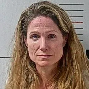 Mugshot of Julie Harding, a woman with long, light brown wavy hair and a white top, against a white wall with height markings.