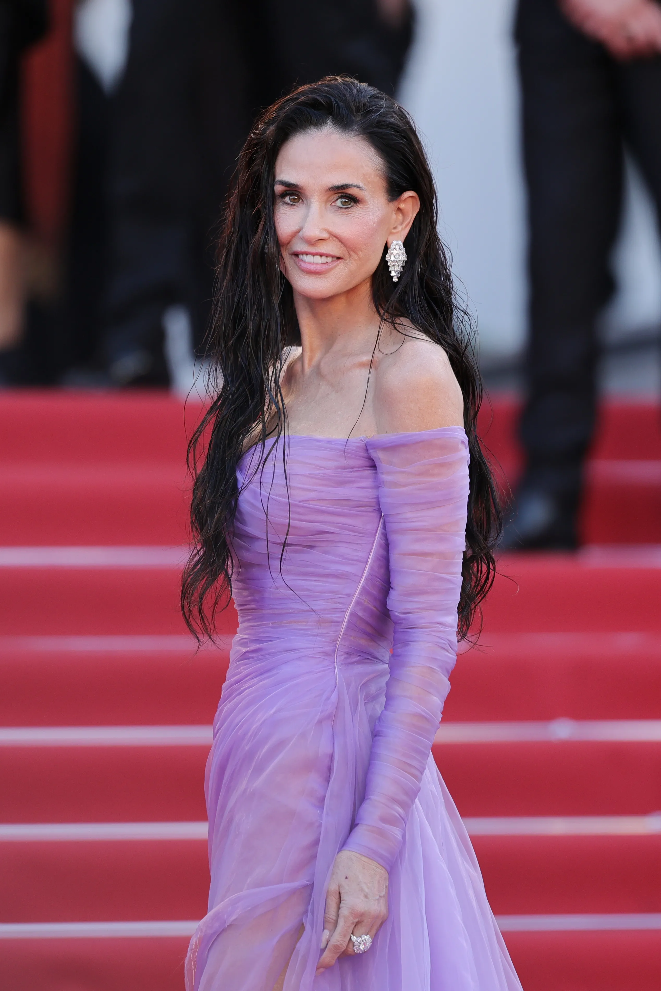 Demi Moore in a purple off-the-shoulder gown and diamond earrings, posing on a red carpet.
