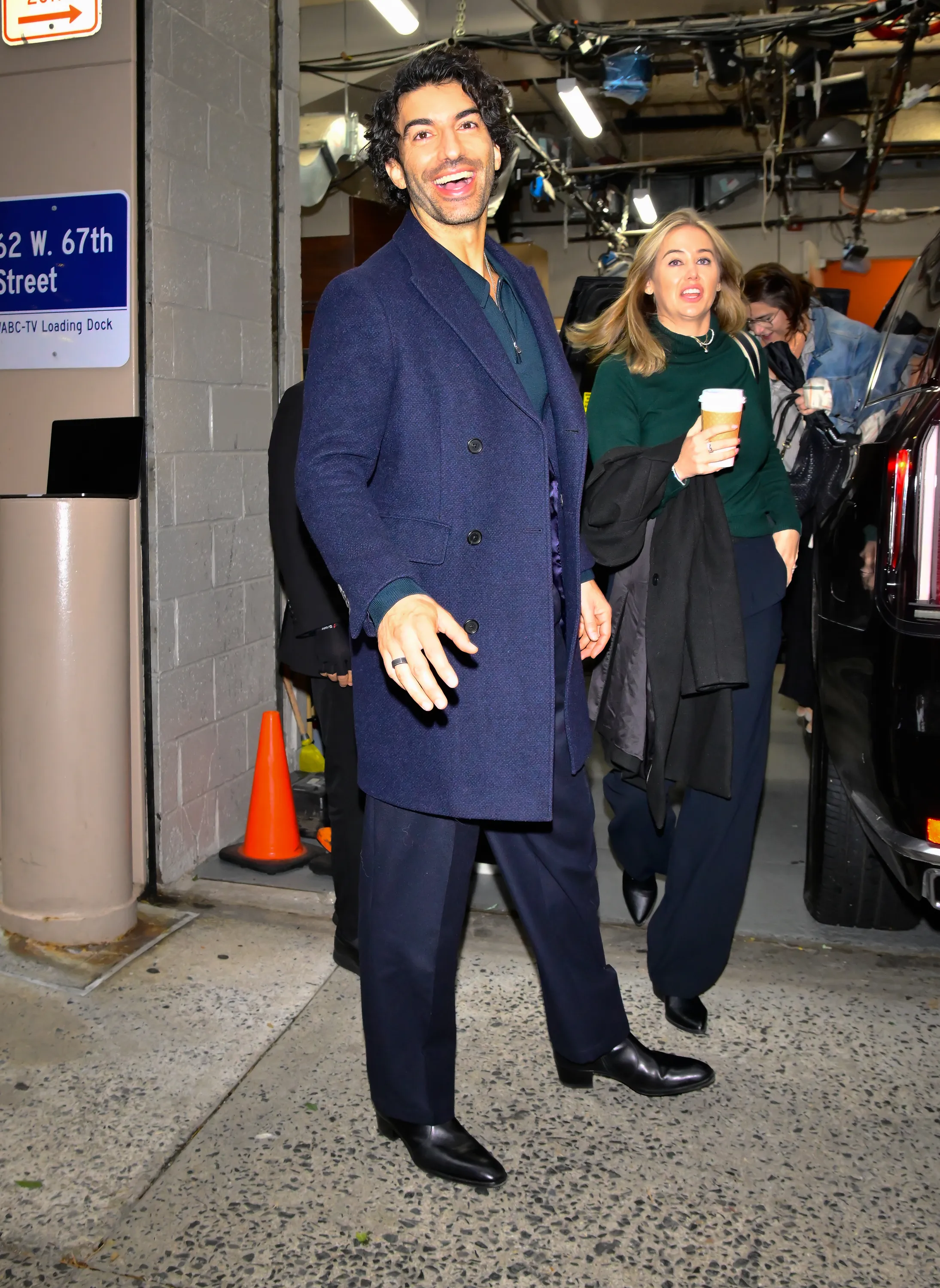 Justin Baldoni in a navy coat and pants, smiling outside ABC Studio.