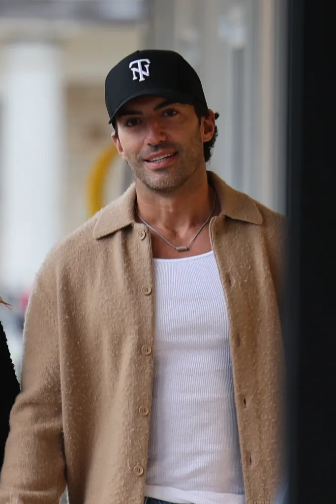 Justin Baldoni smiling, wearing a black cap, a beige open cardigan over a white ribbed tank top, and a silver necklace.