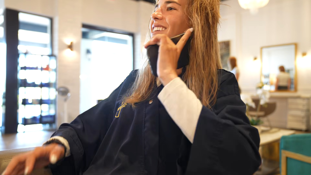 A young woman in a salon robe smiling while talking on a cell phone.