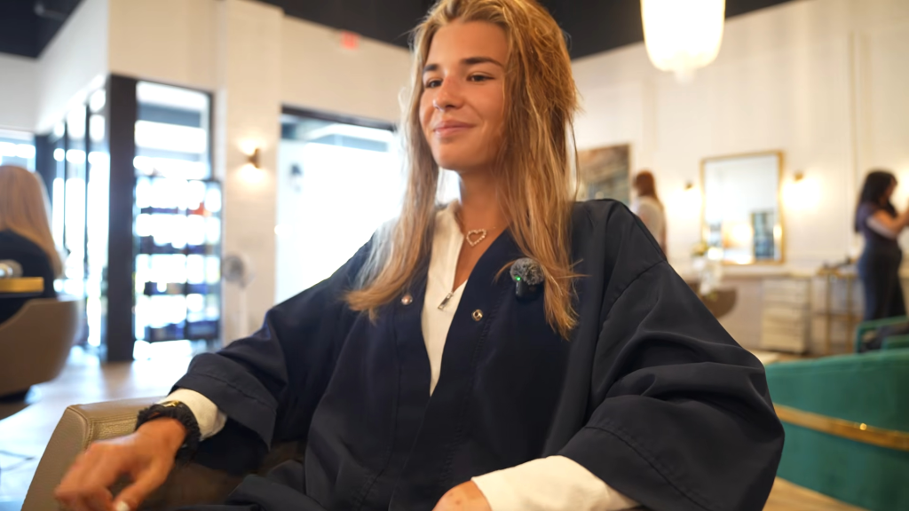 Kai Trump sitting in a salon chair, wearing a black robe over a white shirt and a heart necklace.