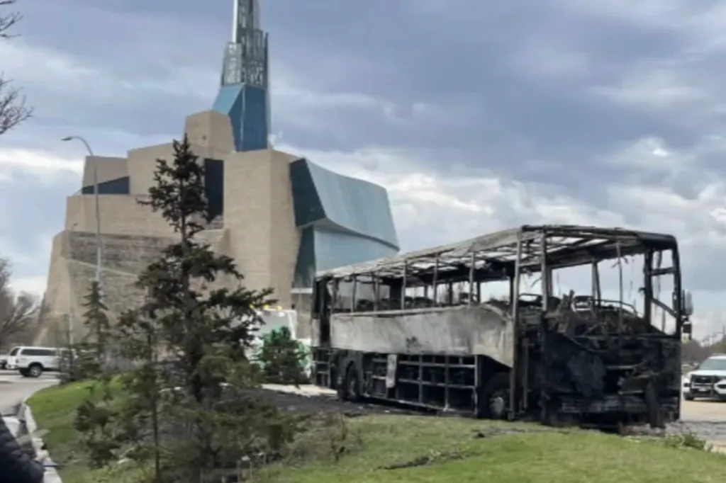 Charred remains of the Kane County Cougars' charter bus on a grassy area next to a road, with a modern building in the background.