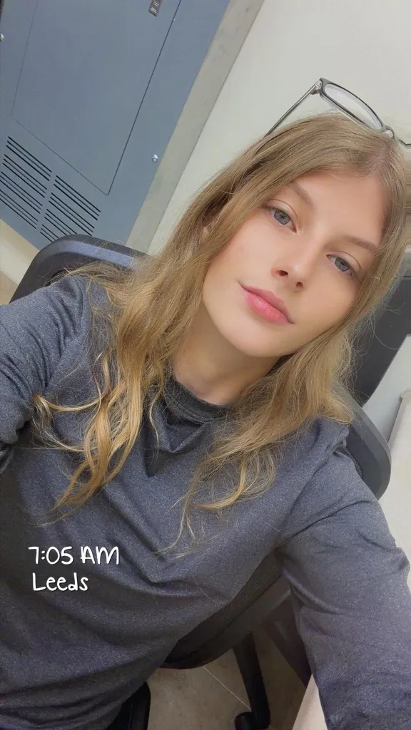 A young woman with long blonde hair and glasses on her head looks at the camera, with