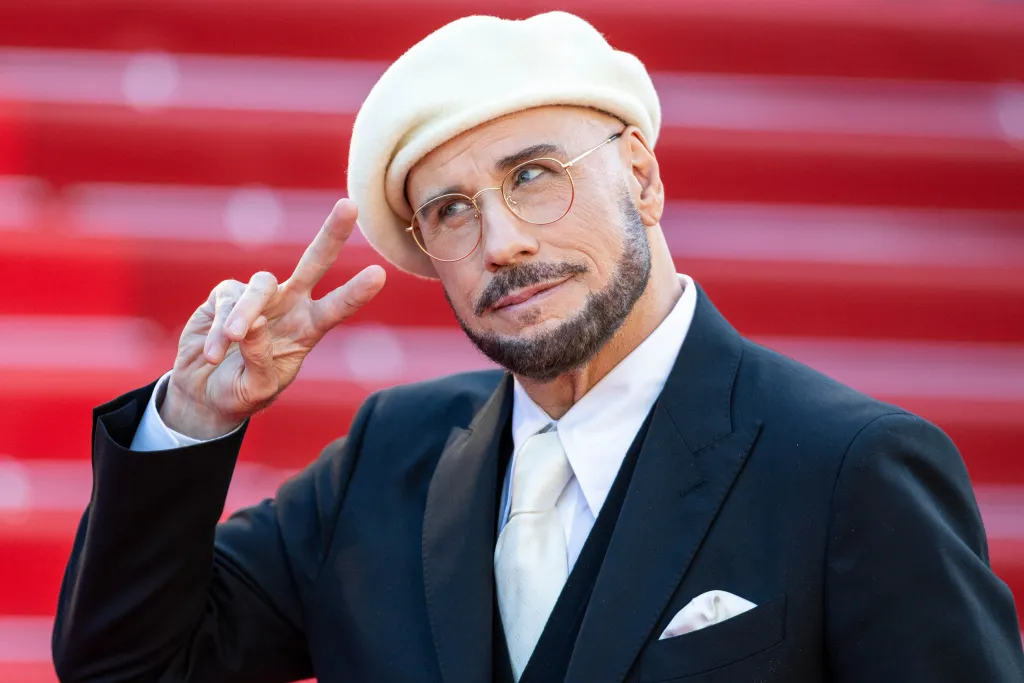 John Travolta wearing a cream beret, black suit, and white tie, making a peace sign on the red carpet at the Cannes Film Festival.
