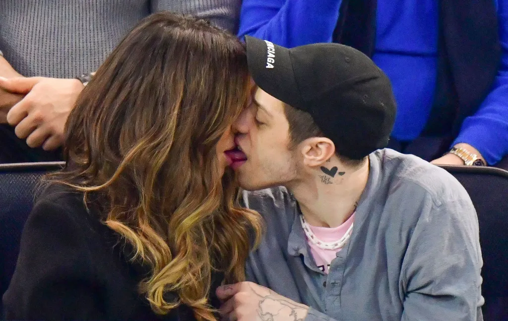 A woman with long wavy brown hair with blonde highlights and a man in a black baseball cap kissing.