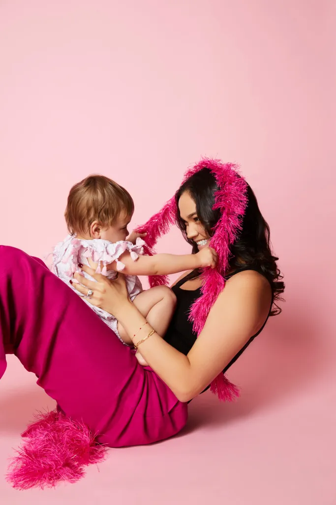 Kate Hernandez in pink pants and black top, holding a baby, with a hot pink feathered scarf around her head.