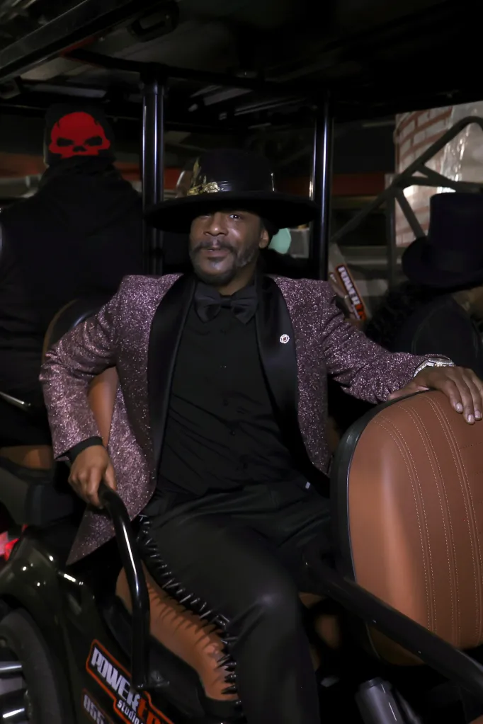 Katt Williams sitting in a golf cart wearing a black hat, a sparkly pink and black tuxedo, and black pants.