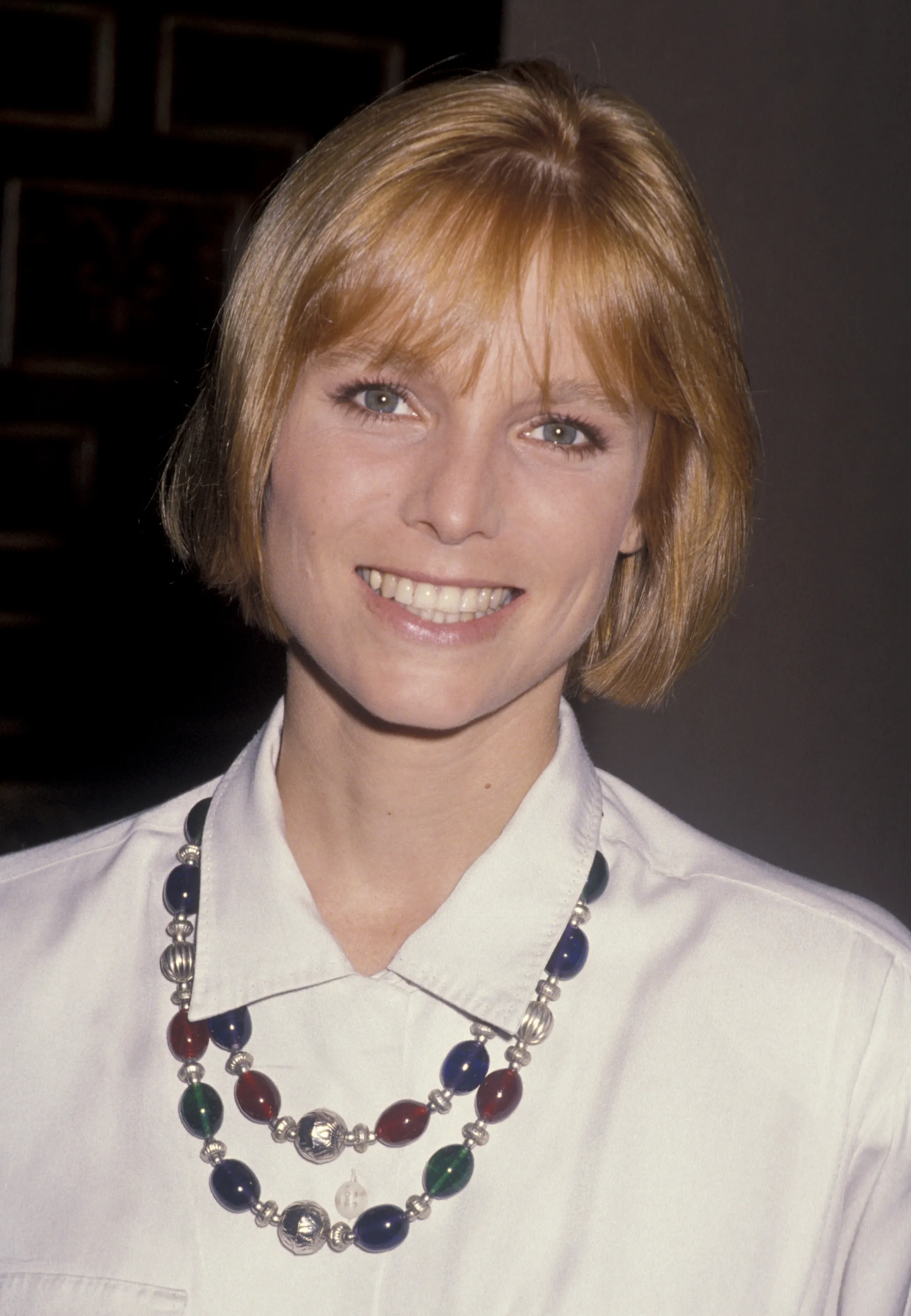 Kelly Curtis smiles wearing a colorful beaded necklace and a white button-up shirt.