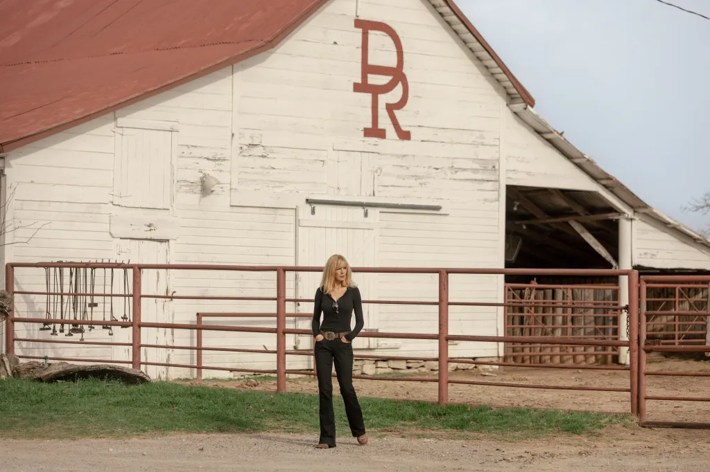 Kelly Reilly as Beth Dutton in black attire with hands in pockets standing in front of a white barn with a red 
