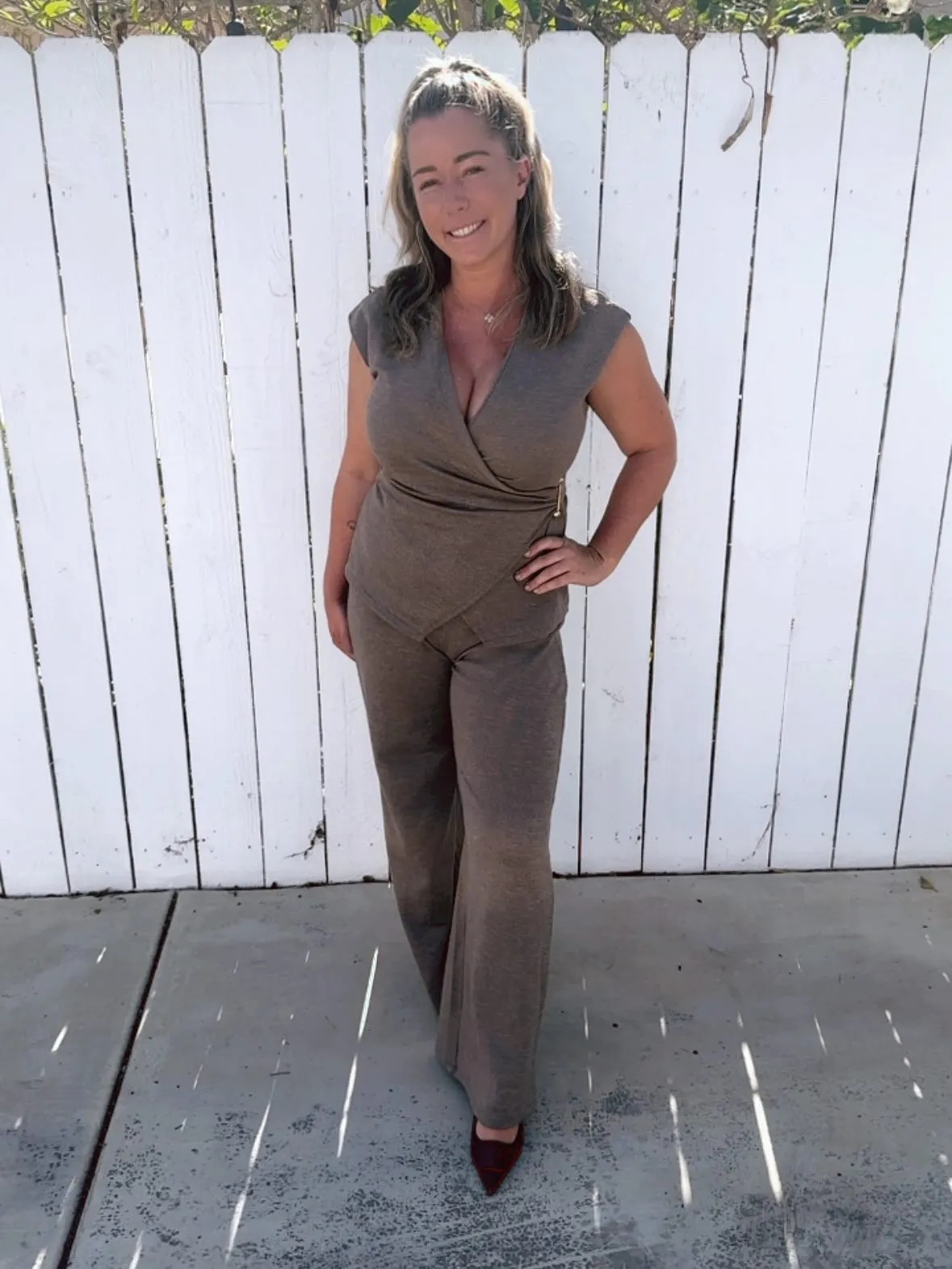 Kendra Wilkinson smiling at the camera, wearing a dark gray two-piece outfit and dark red high heels.