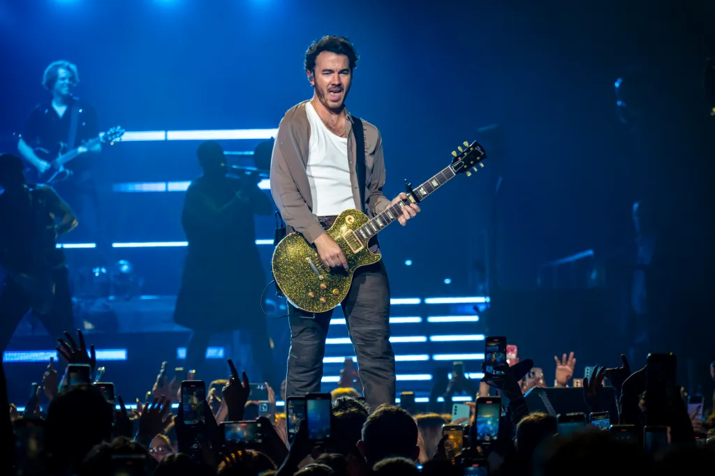 Kevin Jonas performing on stage with a glittery green guitar, facing a crowd holding up cell phones to record.