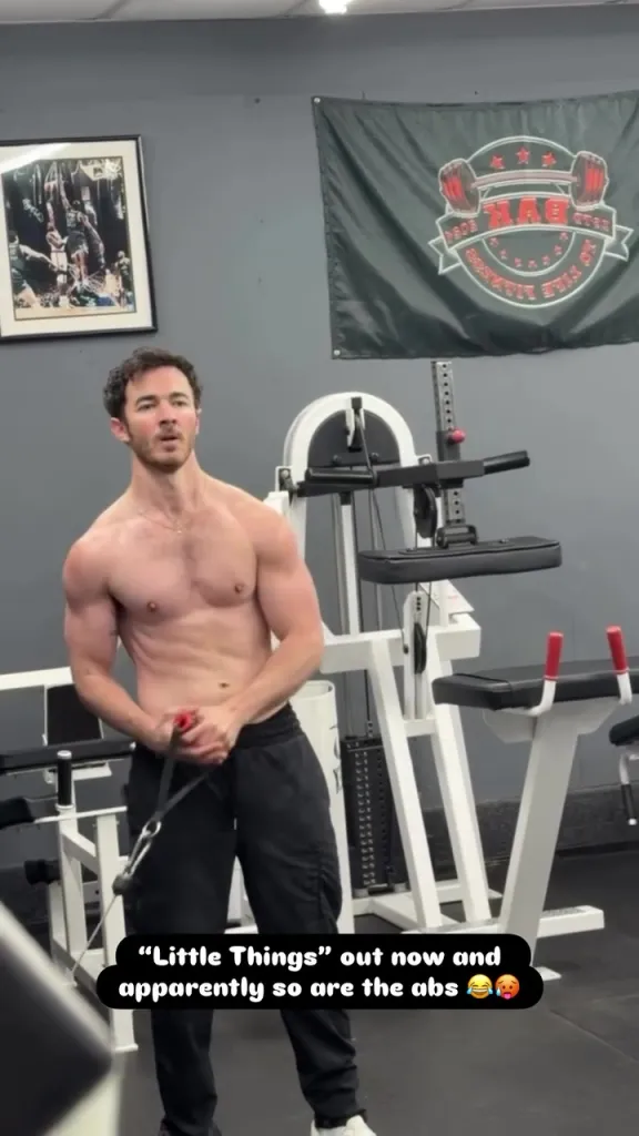 Kevin Jonas posing shirtless in a gym with visible abs, holding a resistance band.