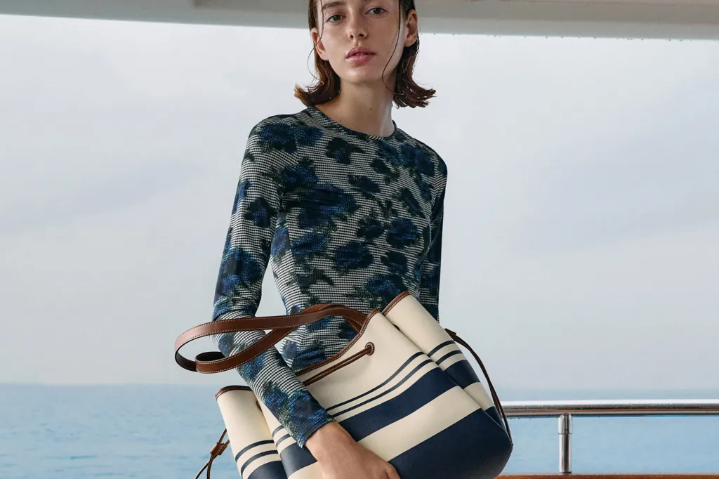 A woman in a blue and black patterned shirt holds a striped cream and navy tote bag.