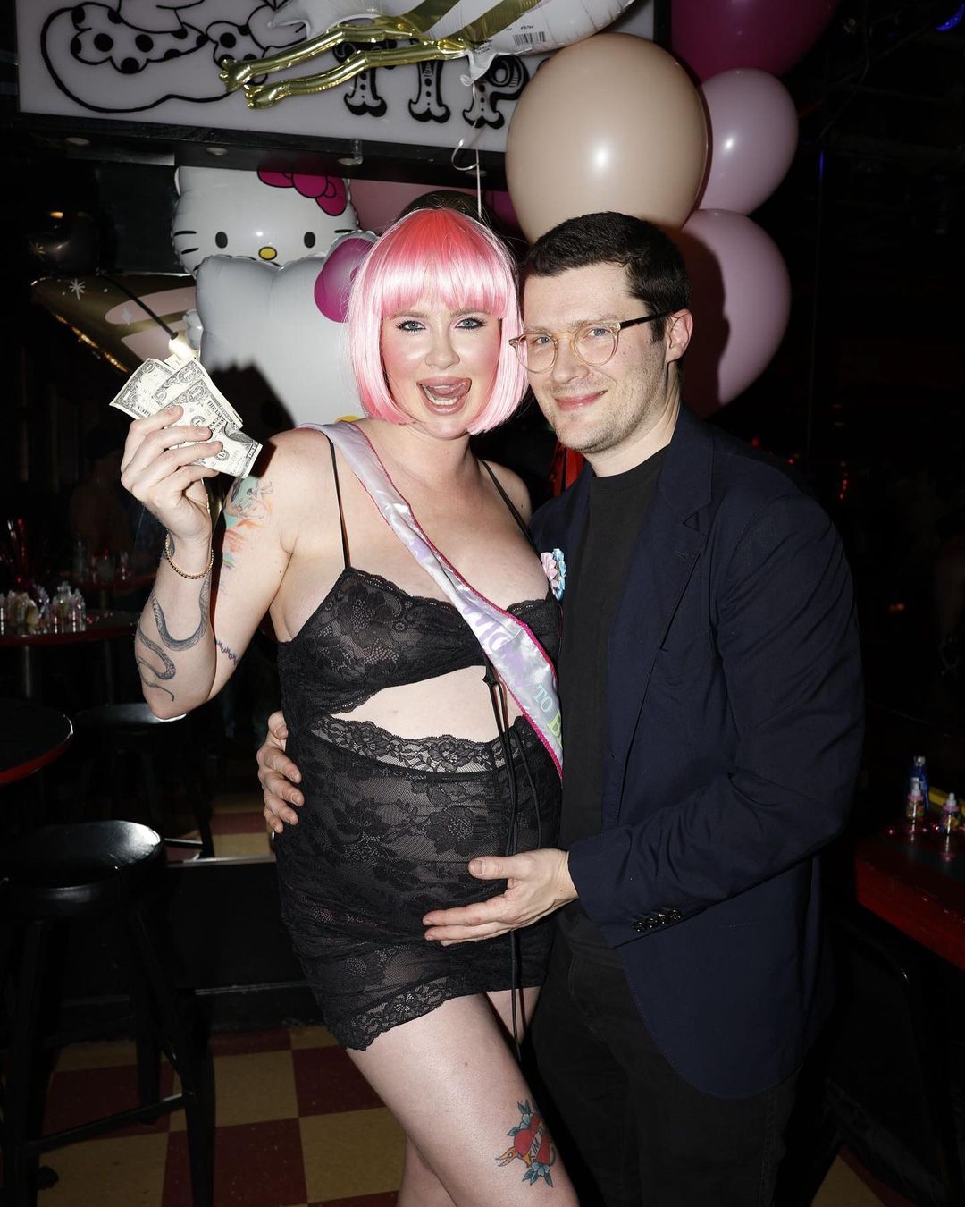 Ireland Baldwin, wearing a pink wig, holding money, and boyfriend RAC, holding her pregnant belly at her baby shower.