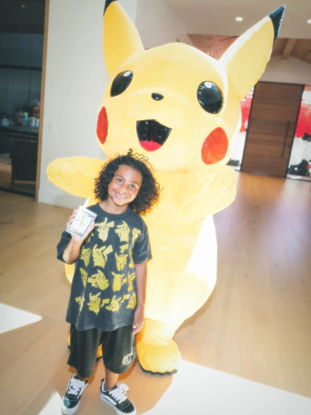 Psalm West holding a Pokémon card with a large Pikachu mascot behind him.