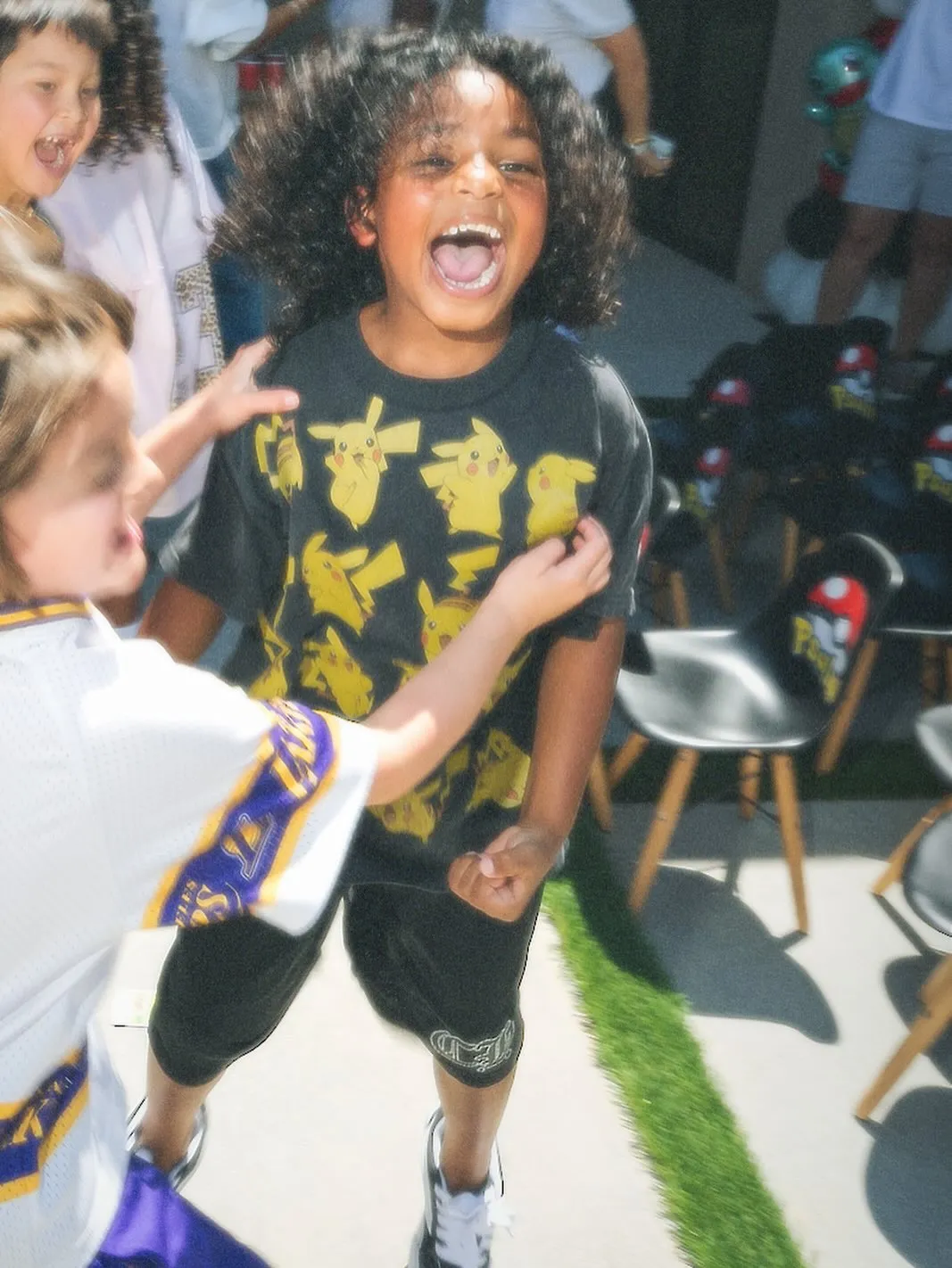 A child with curly hair wearing a Pikachu shirt smiles excitedly at a Pokémon-themed birthday party.