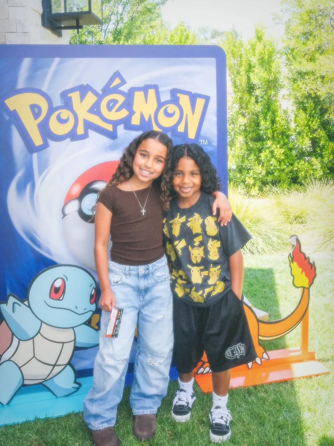 Two children posing in front of a giant Pokémon logo with cutouts of Squirtle and Charizard.