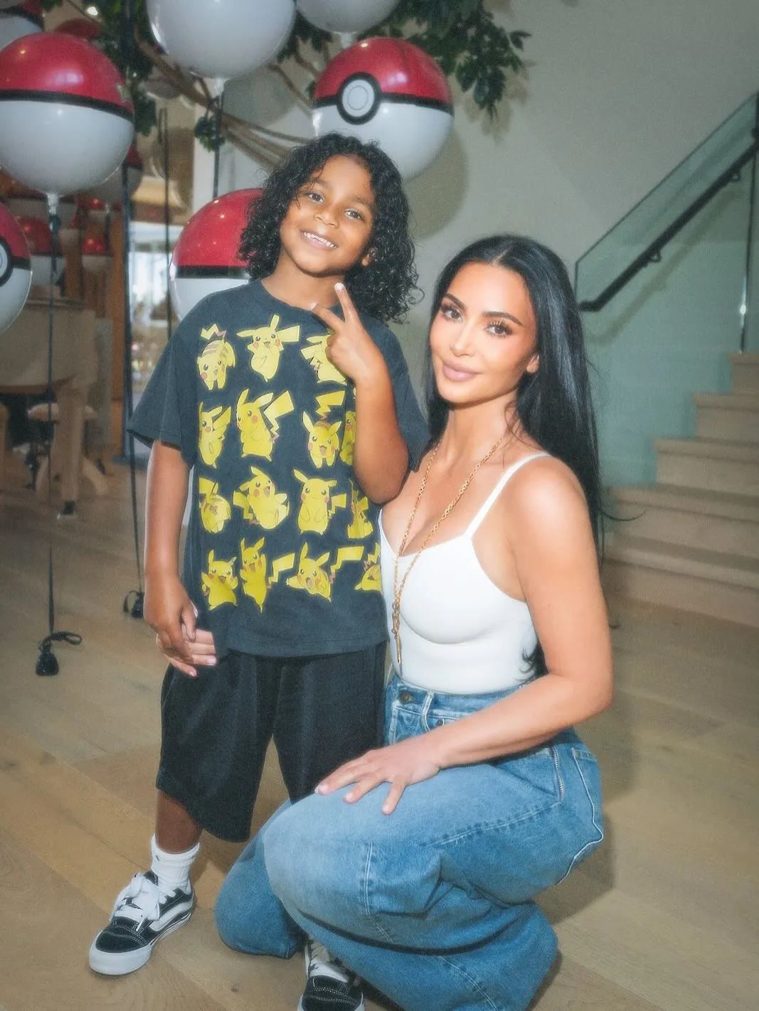 Kim Kardashian kneels next to Psalm, both smiling, with Pokémon balloons in the background.