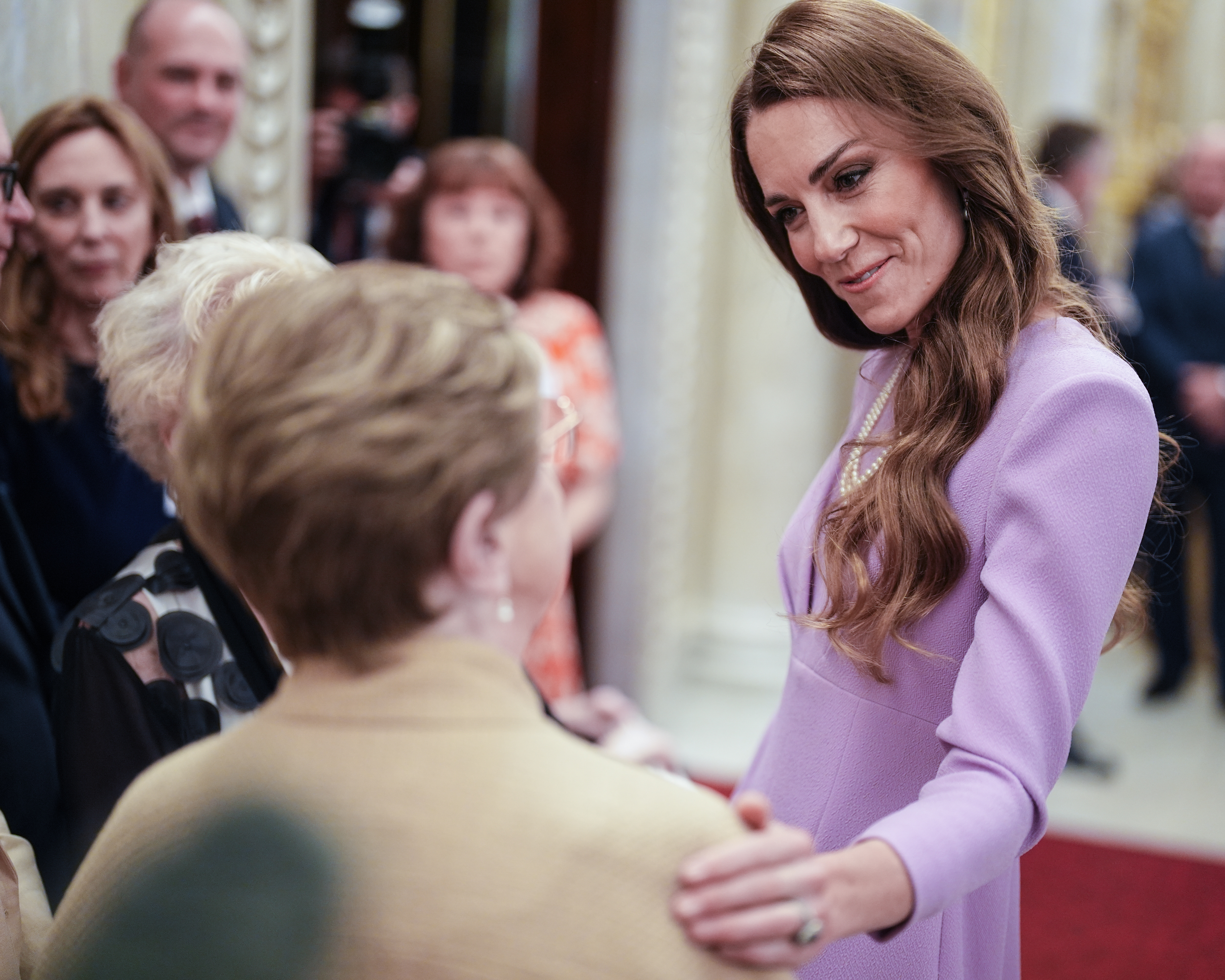 Catherine, Princess of Wales, in a lilac dress, smiles while greeting a woman at Buckingham Palace.