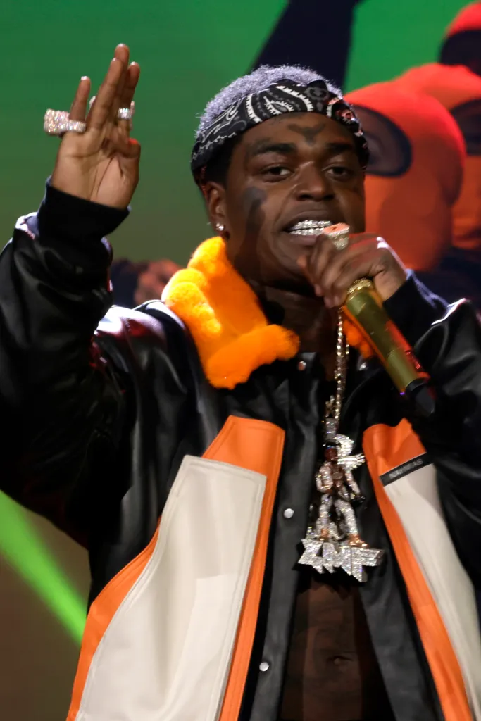 Kodak Black performing onstage in a black and orange jacket, gold grill, and a diamond necklace.