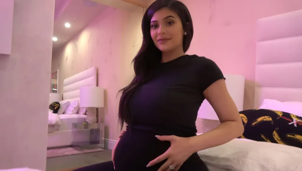 Kylie Jenner sitting in a room with pink lighting, cradling her baby bump.