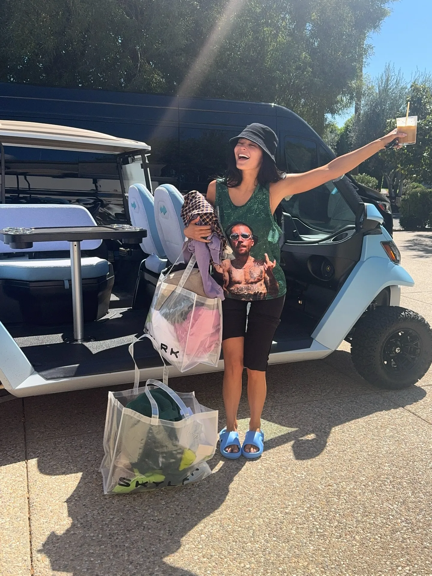 Kylie Jenner in a green tank top, black shorts, and blue slides, holding a drink and bags, next to a golf cart.