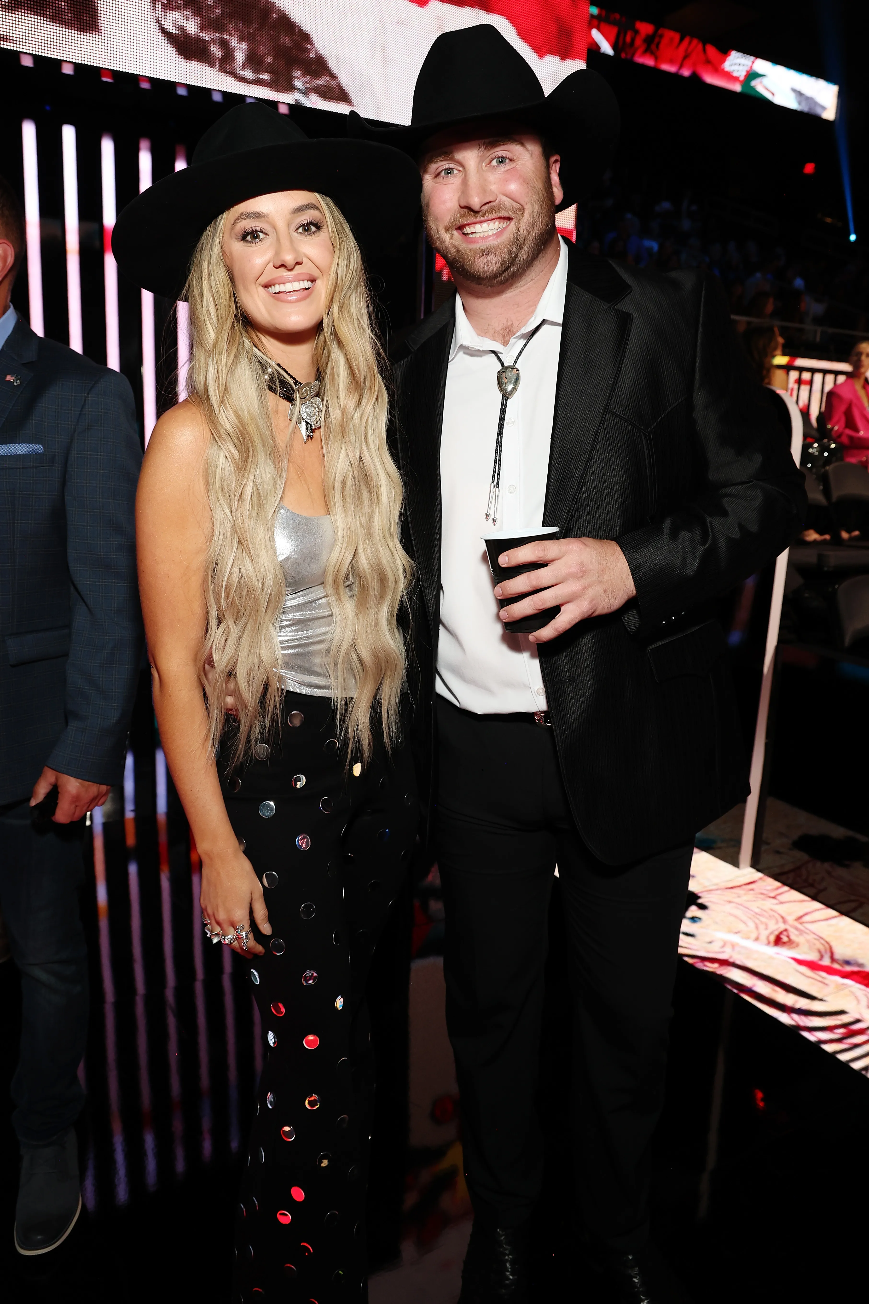 Lainey Wilson and Devlin Hodges pose wearing cowboy hats, Wilson in a silver top and polka dot pants, Hodges in a bolo tie and black suit.