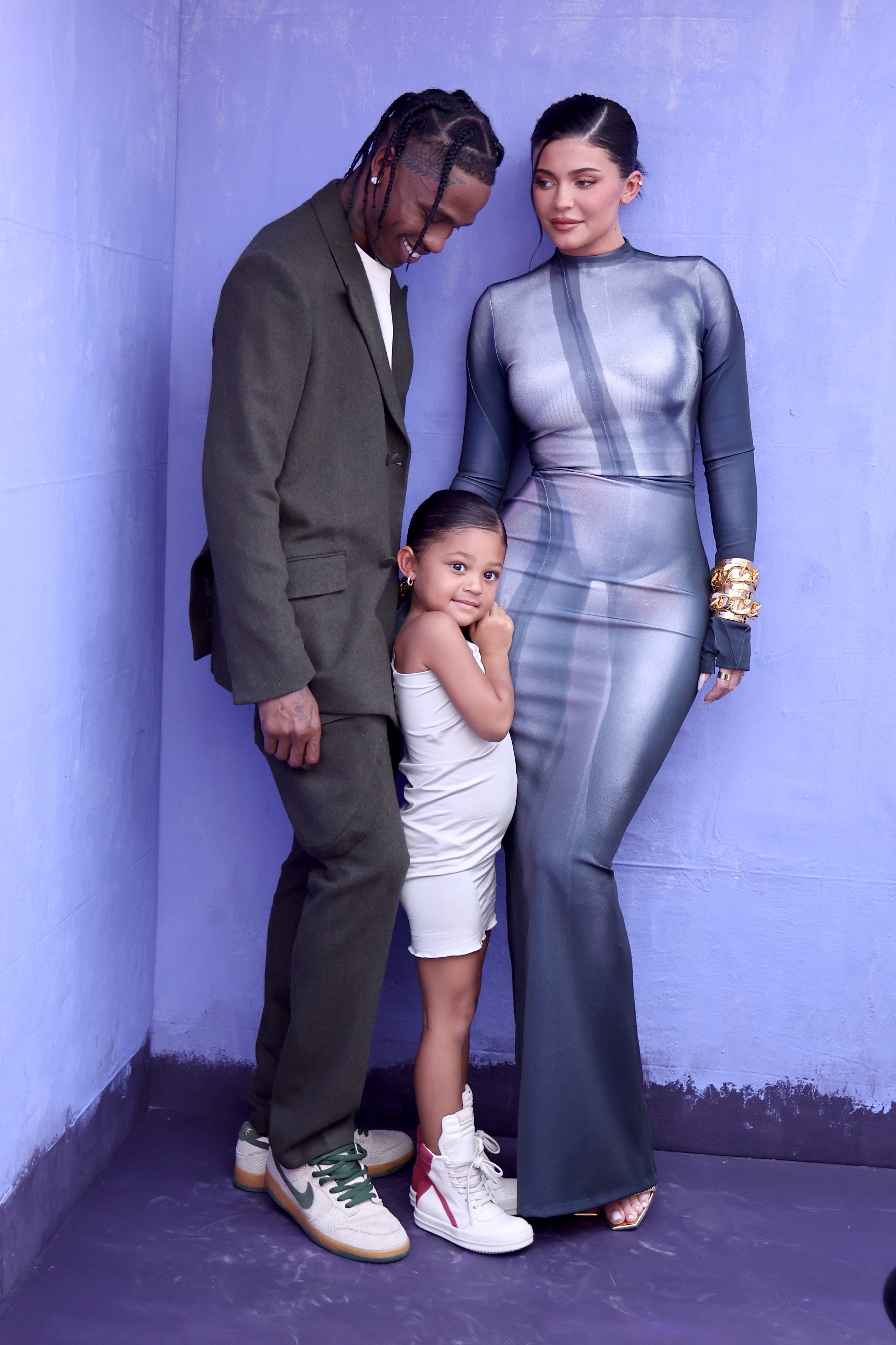 Travis Scott, Stormi Webster, and Kylie Jenner standing against a lavender wall.