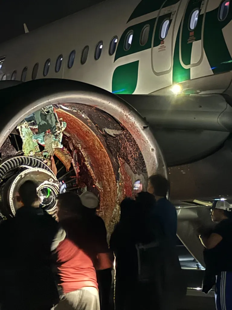 A damaged Frontier Airlines jet engine, with visible blood and debris, is inspected by people at night.