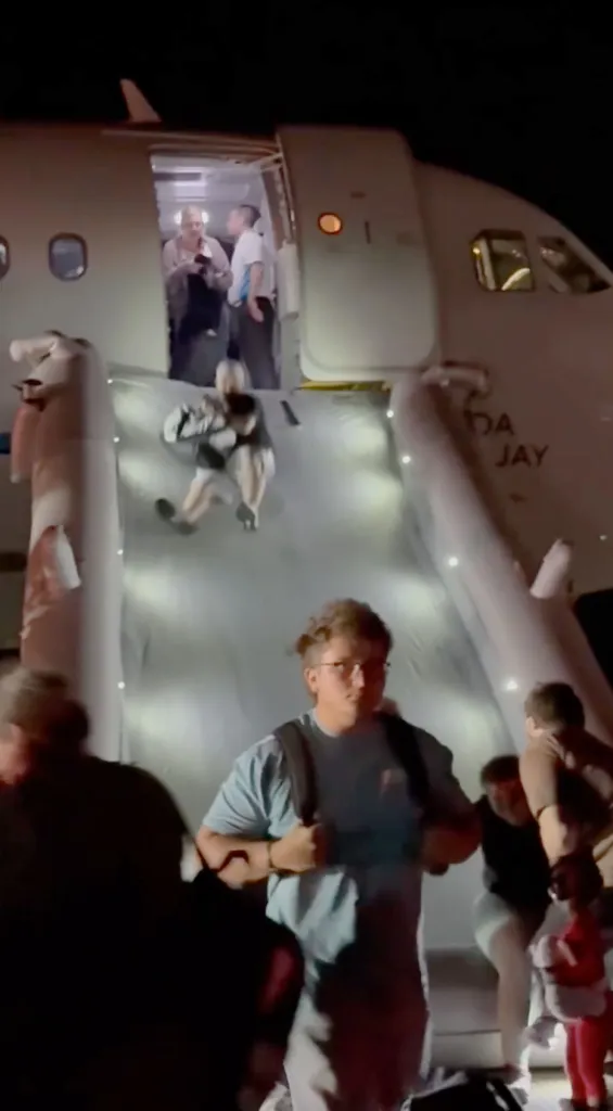 Passengers slide down an emergency inflatable chute from an airplane at night.