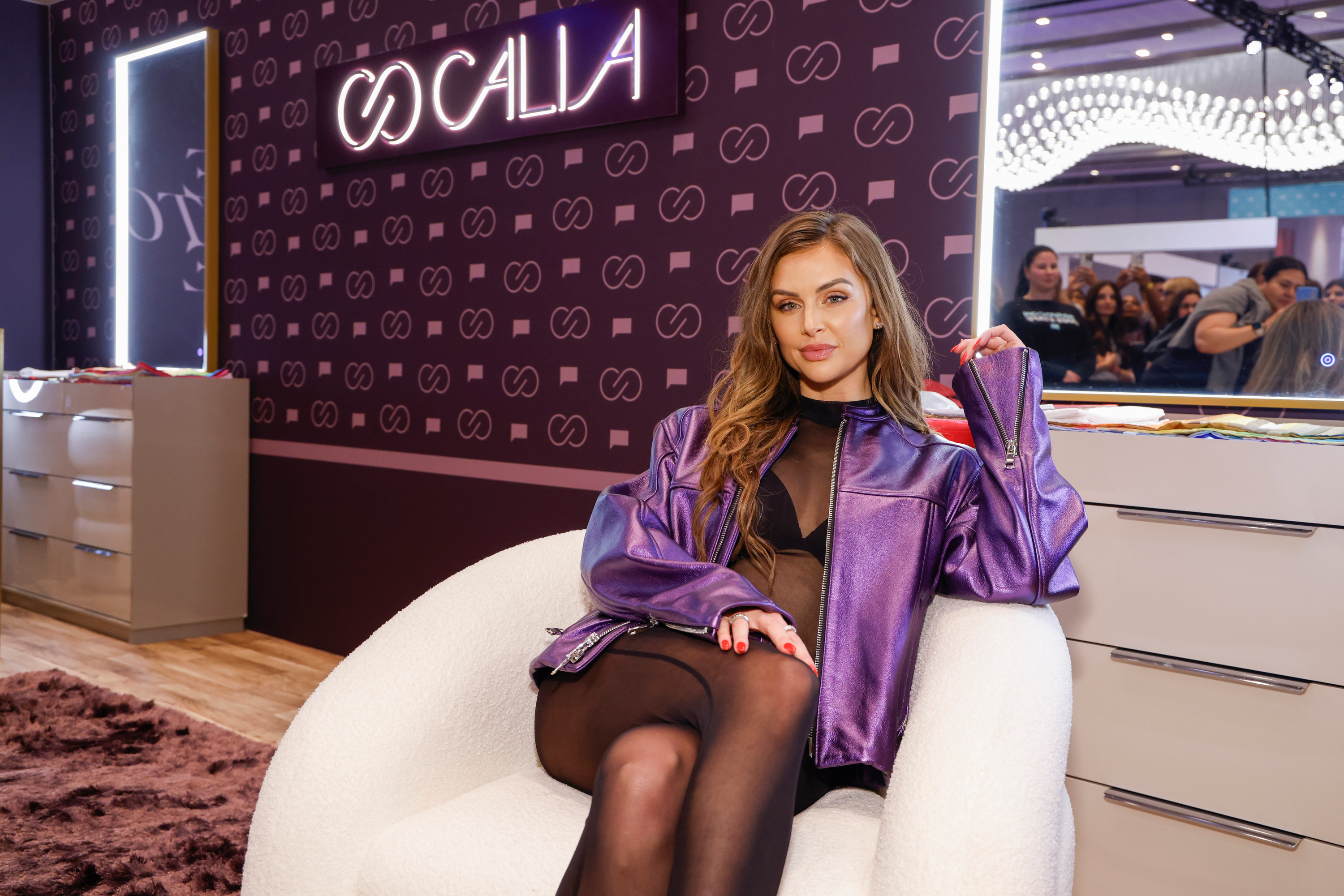 Lala Kent posing in a purple jacket and black tights while sitting in a white armchair.