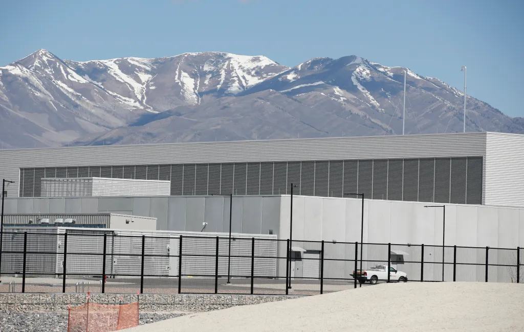 A long gray building with large air vents runs along the base of snow-capped mountains.