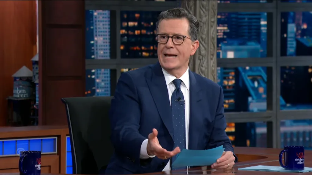 Stephen Colbert in a suit and tie, seated at his desk with city lights in the background.