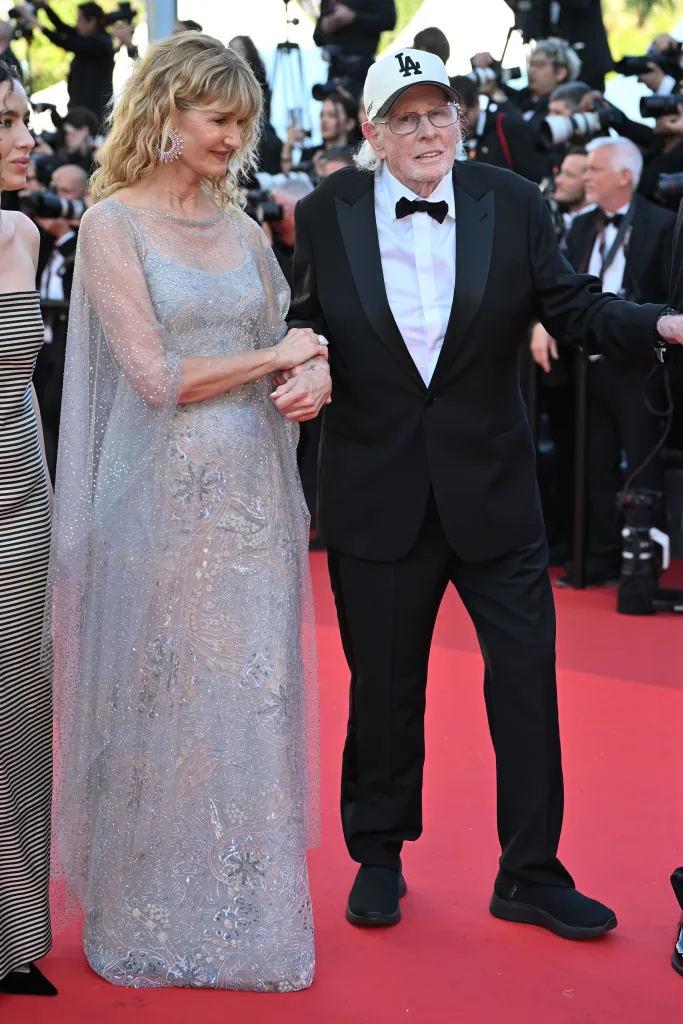 Laura Dern in a silver gown and Bruce Dern in a tuxedo with a white LA Dodgers cap on a red carpet.